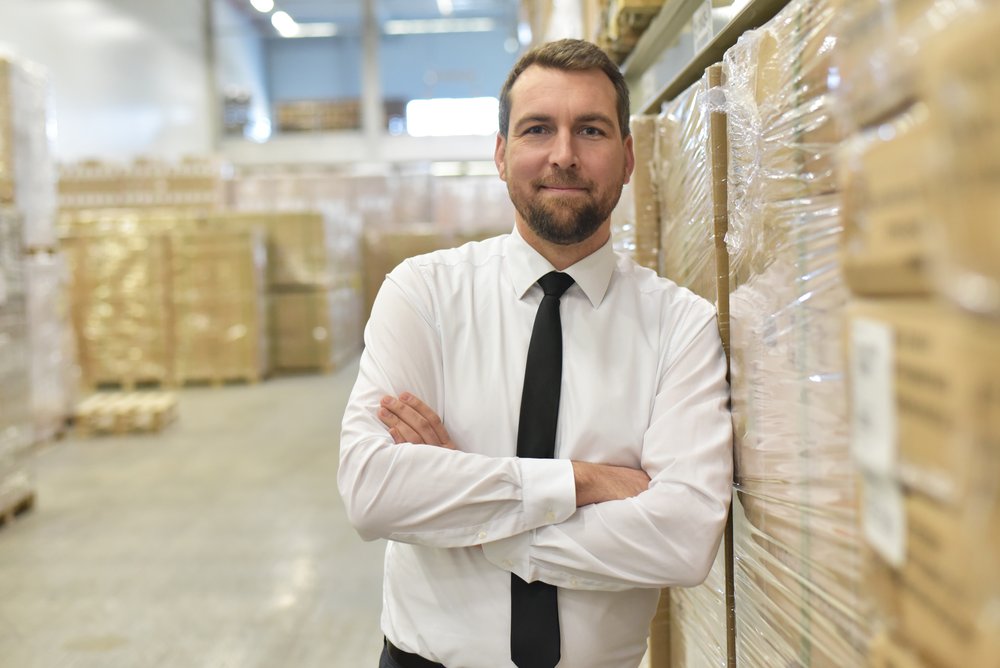 Man with a beard in a white suit with a black tie on. He is stood next to a pallet of boxes in a warehouse.
