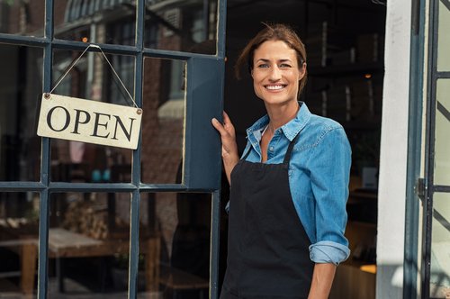 Small female business owner wearing a denim shirt stood outside a door that reads 'open'.