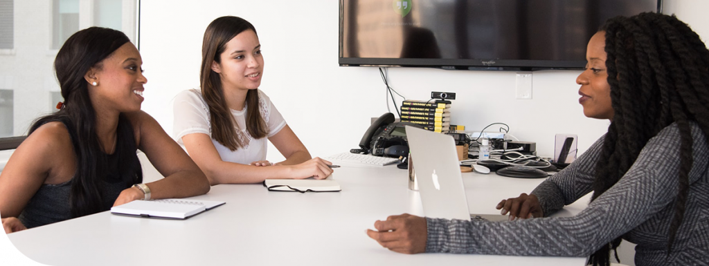 Three women sat at a meeting room table. Two have notebooks and 1 has a laptop in front of them.