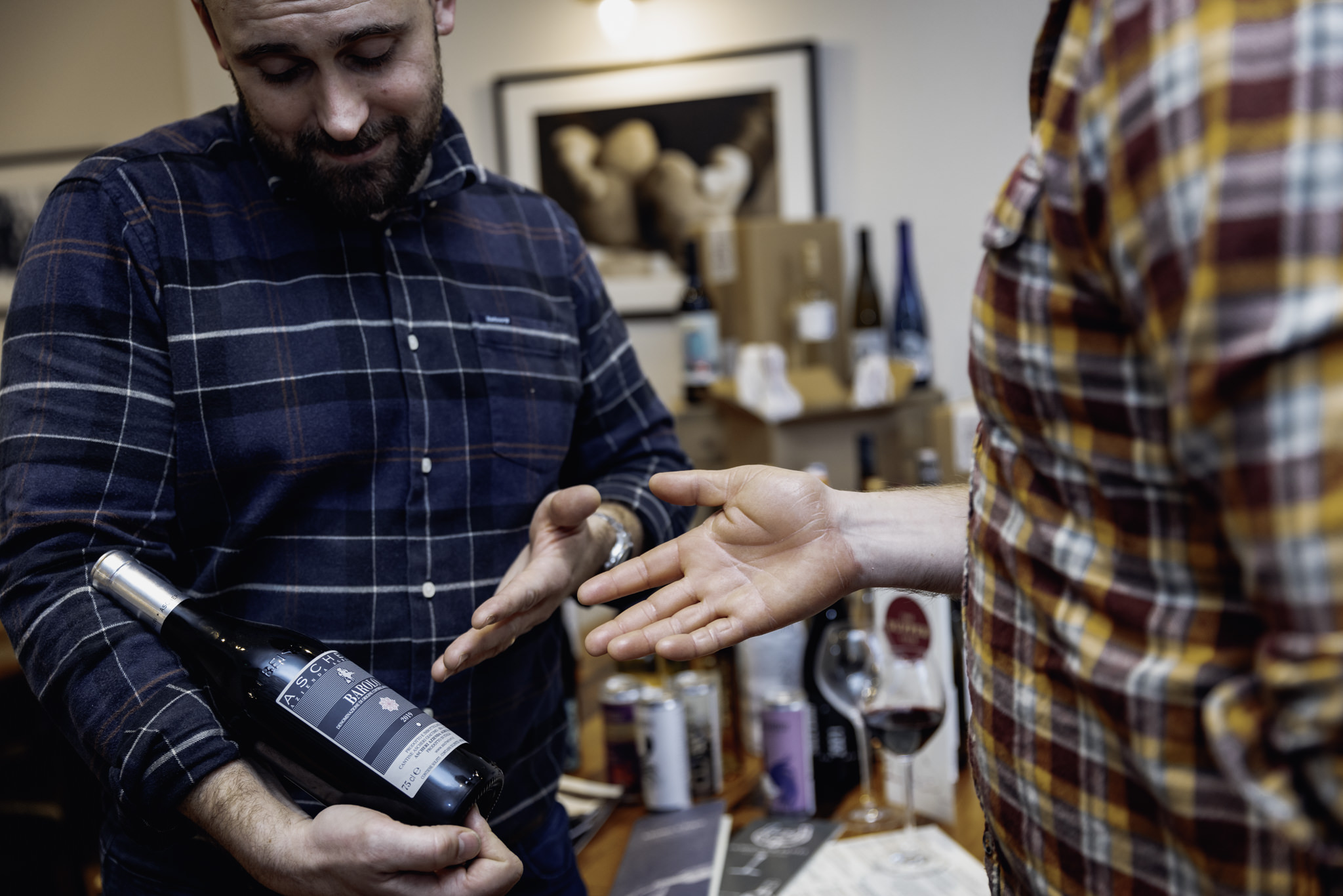 A pair of men talk in a wine shop as one holds a bottle of red wine