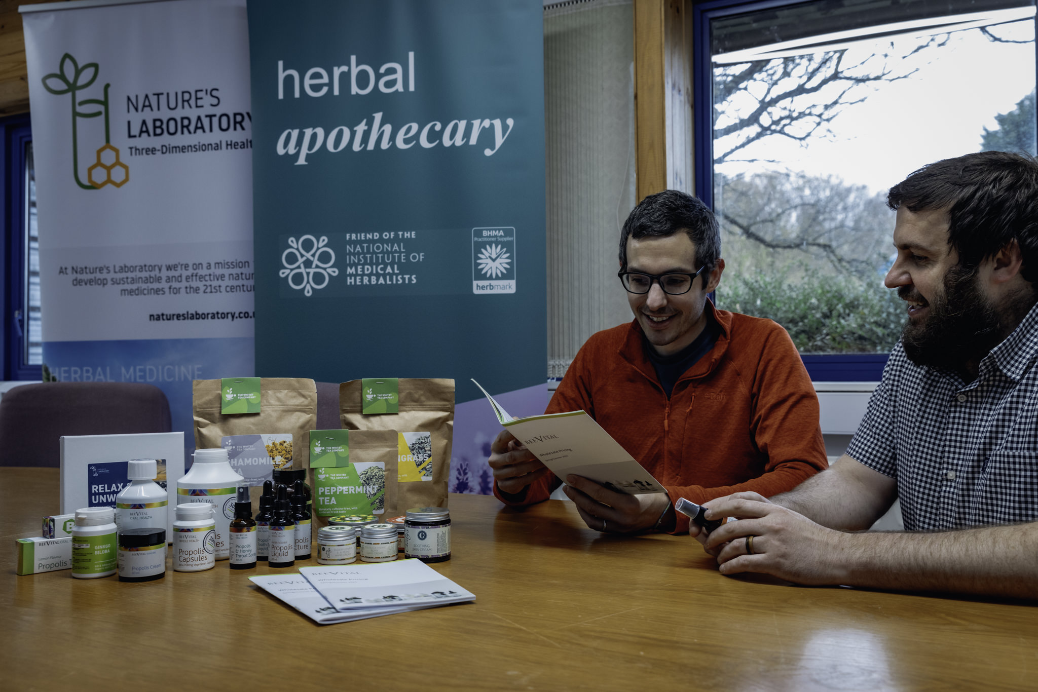 Two men sit at a table with some herbal supplements, behind them are two banners for herbal supplements